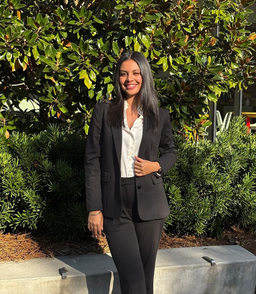 Simar in a black suit and white shirt, standing in front of magnolia trees