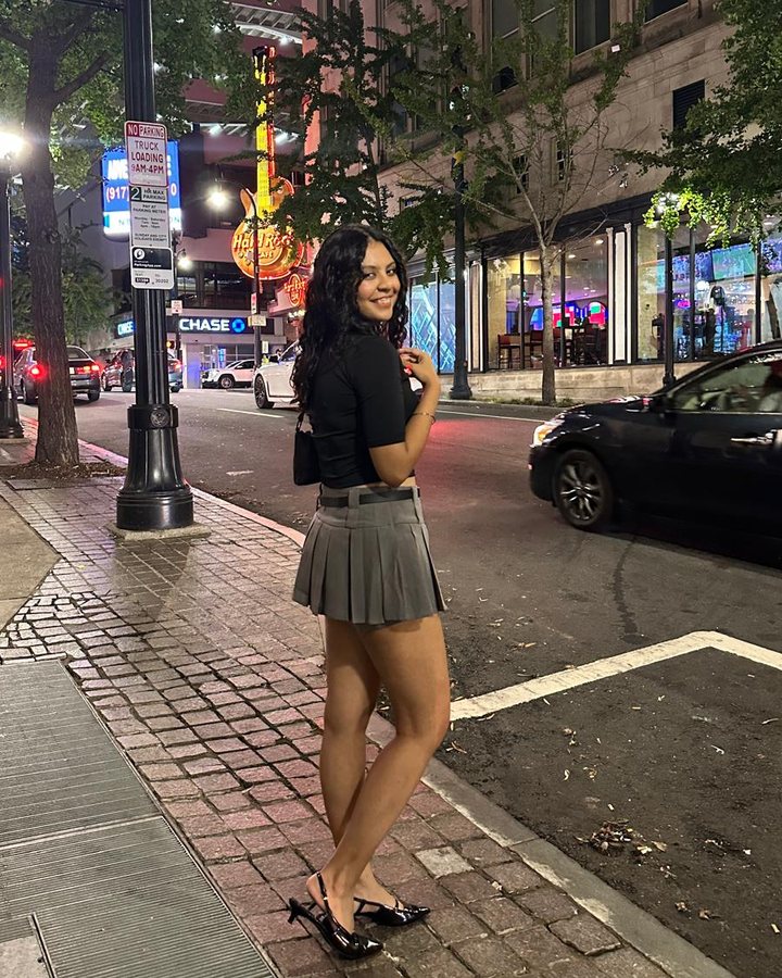 Simar on a city street at night in a black top and grey pleated skirt